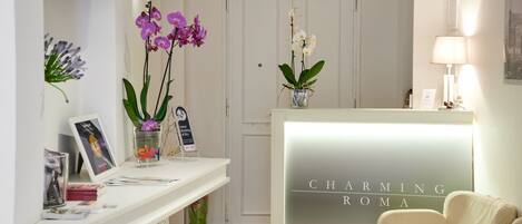 Un espace d’accueil avec un comptoir de réception portant l’inscription « CHARMING ROMA », un mur blanc orné d’un tableau encadré, un vase contenant des orchidées violettes et une petite table sur laquelle sont posés un panneau et une photographie encadrée.