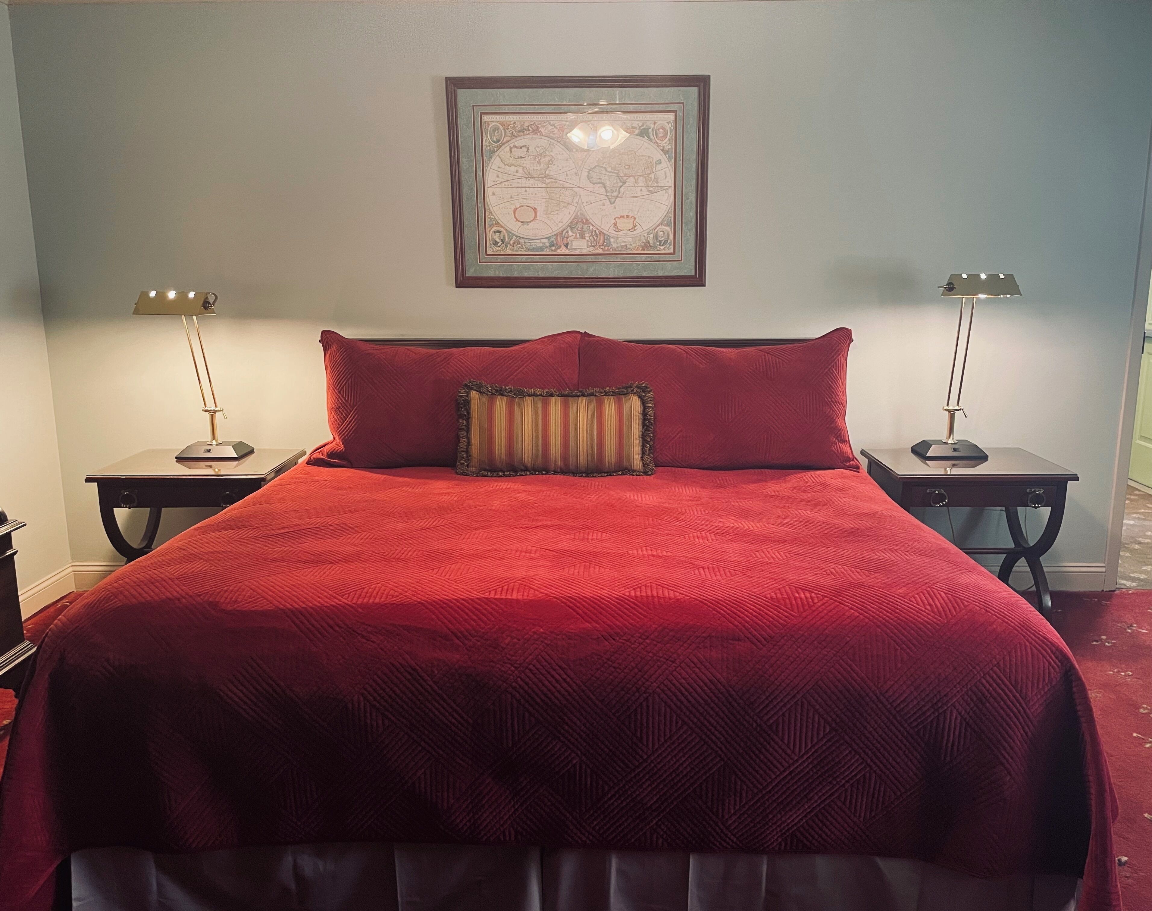 A neatly made bed with a red quilt, two bedside lamps, and a framed map on the wall.