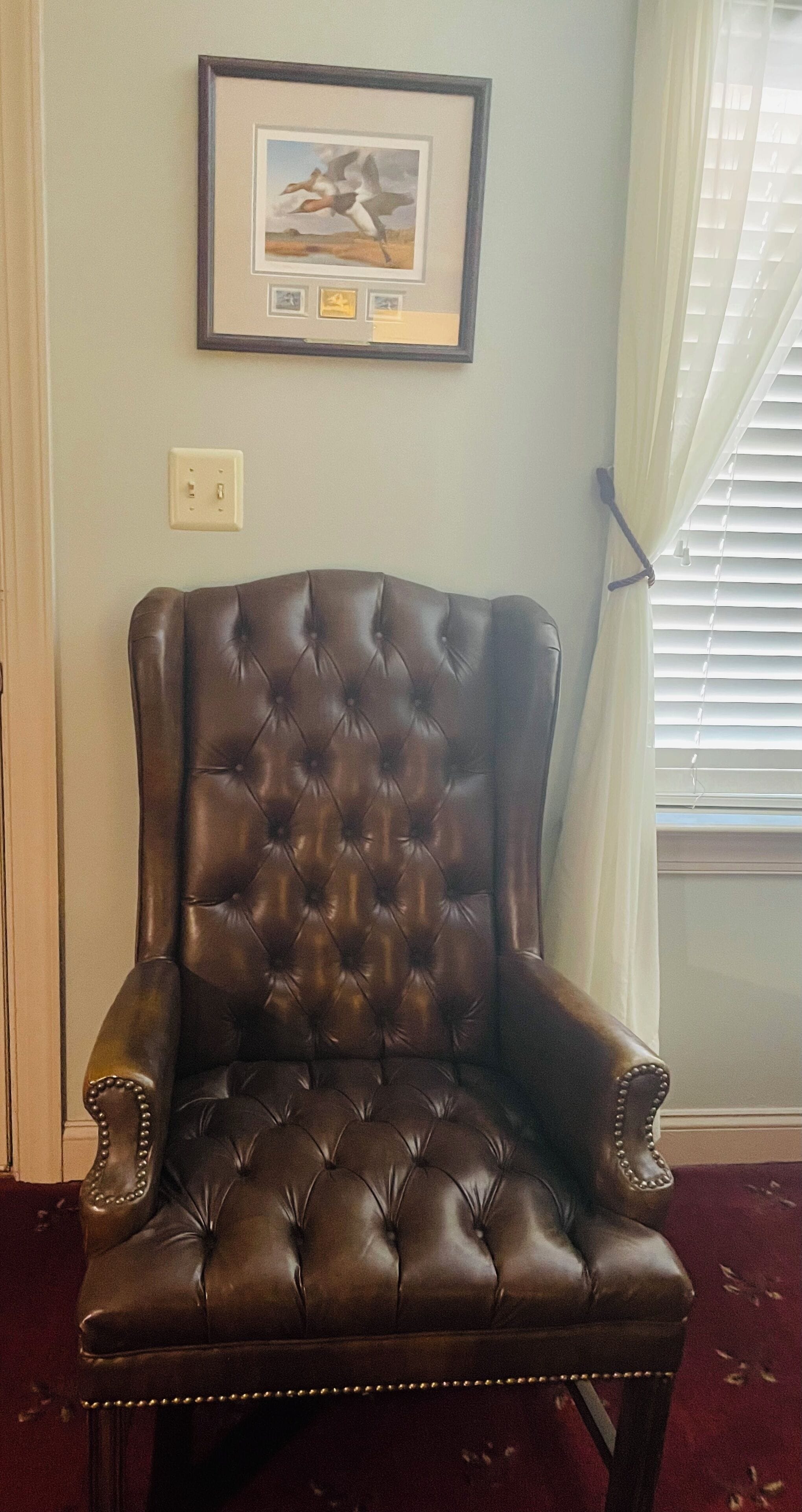 Un sillón de cuero capitonado con asiento y respaldo acolchados, ubicado en una habitación con un cuadro enmarcado en la pared y una ventana con persianas.