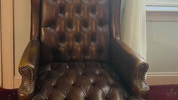 Un sillón de cuero capitonado con asiento y respaldo acolchados, ubicado en una habitación con un cuadro enmarcado en la pared y una ventana con persianas.