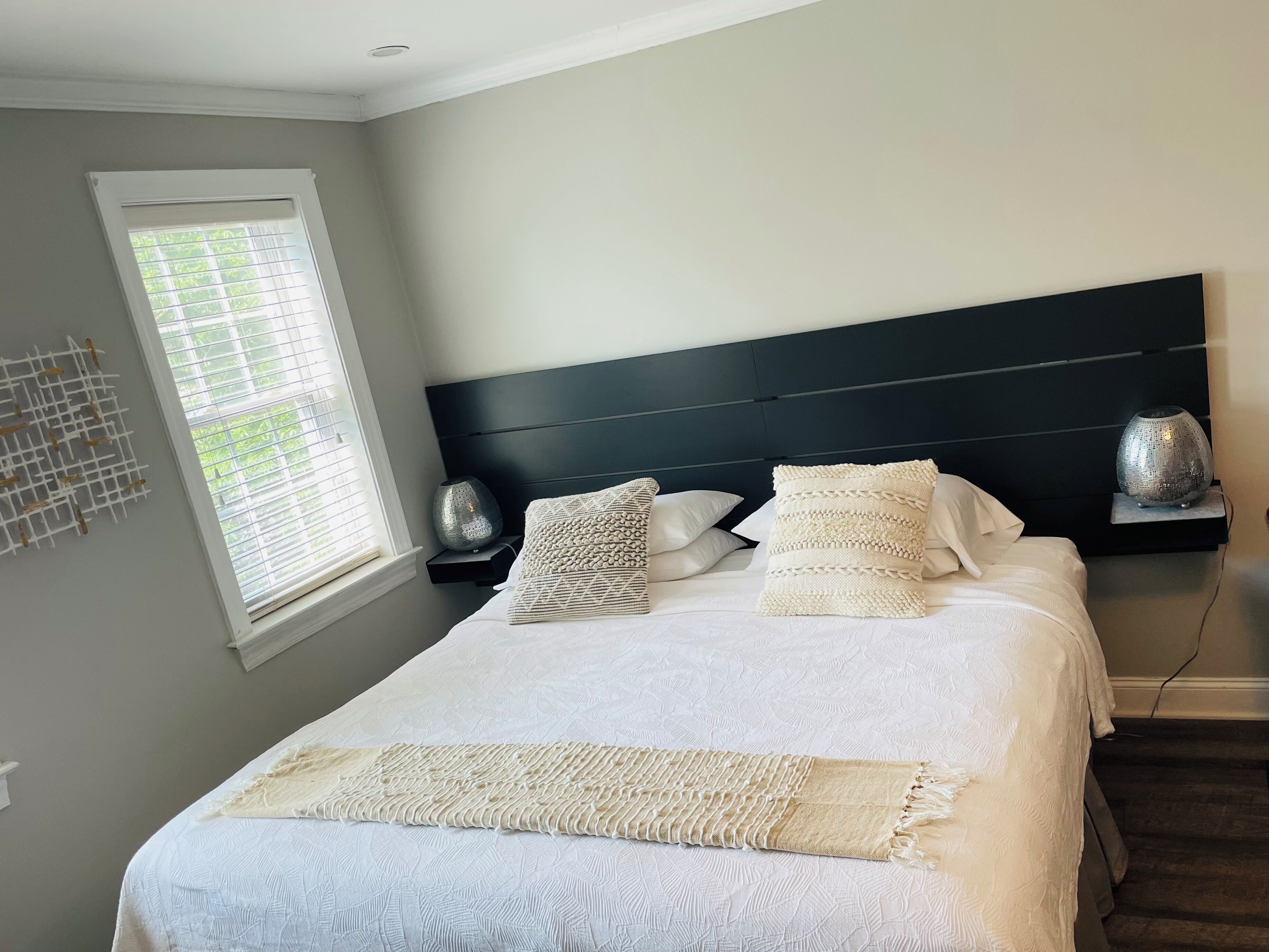 A neatly made bed with a white comforter and decorative pillows, a bedside table with a lamp, and a window with blinds.