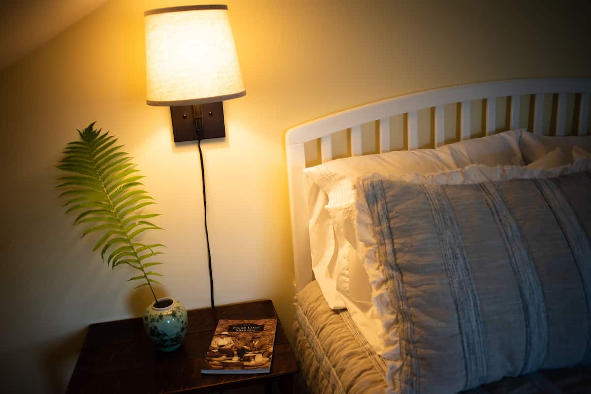 A neatly made bed with white bedding and a headboard, a bedside lamp, a small table with a book and a decorative plant.