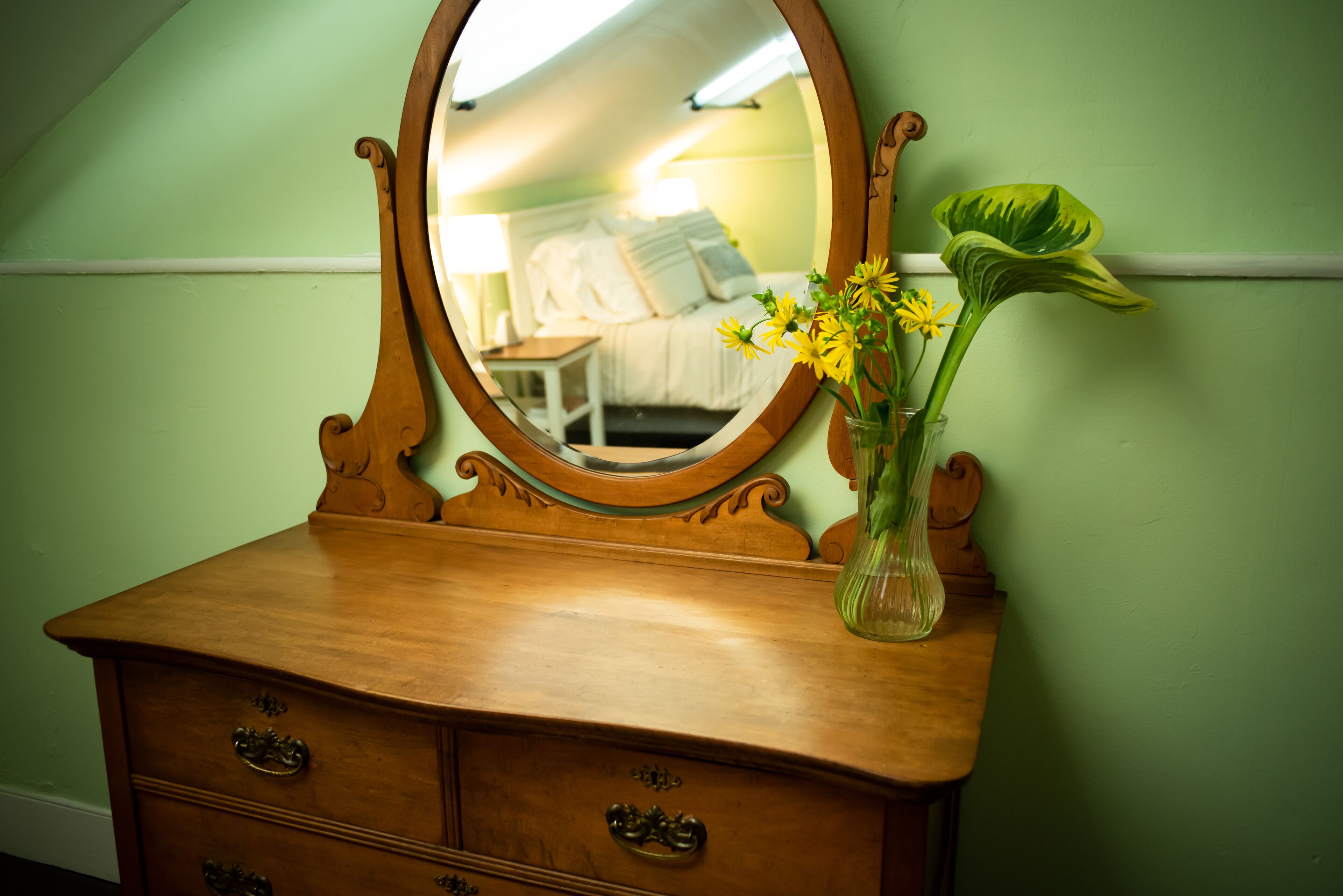 A wooden dresser with a mirror, a vase with yellow flowers, and a green wall.