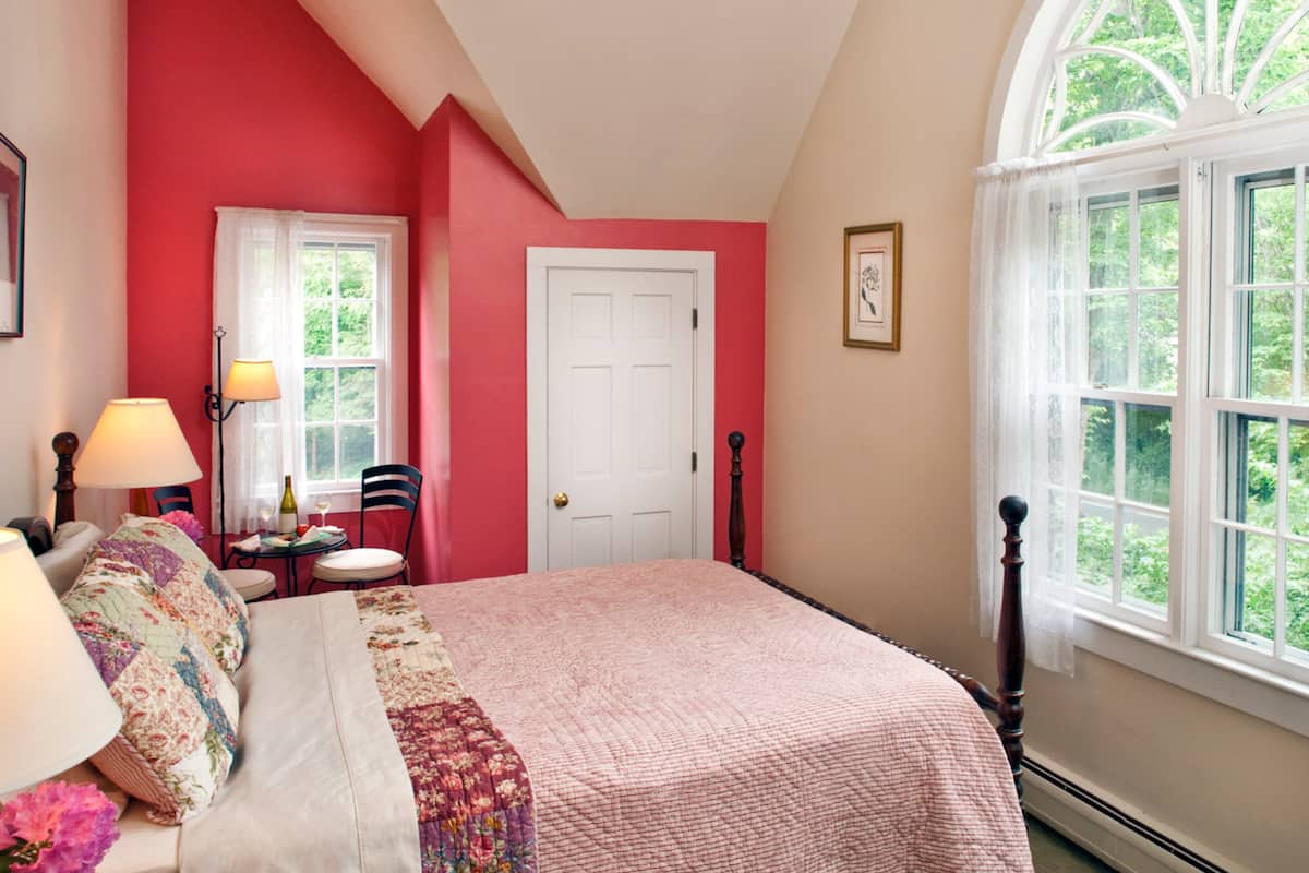A bedroom with a large bed, a window with a view of greenery, a door, and a small table with a lamp.