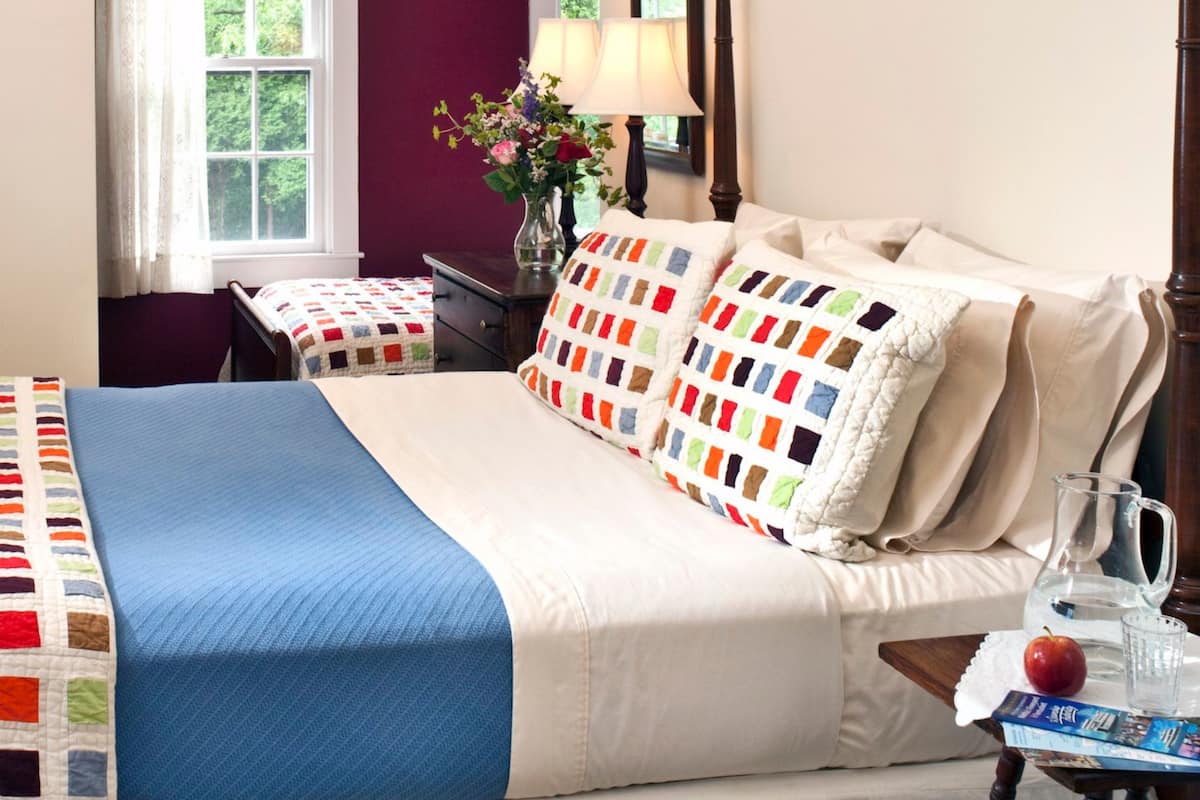 A neatly made bed with a blue blanket and a colorful quilt, a bedside table with a lamp, a vase with flowers, and a window with curtains.