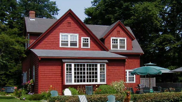 Una casa rossa con un'ampia finestra sul davanti, un portico coperto e due poltrone da giardino Adirondack verdi.