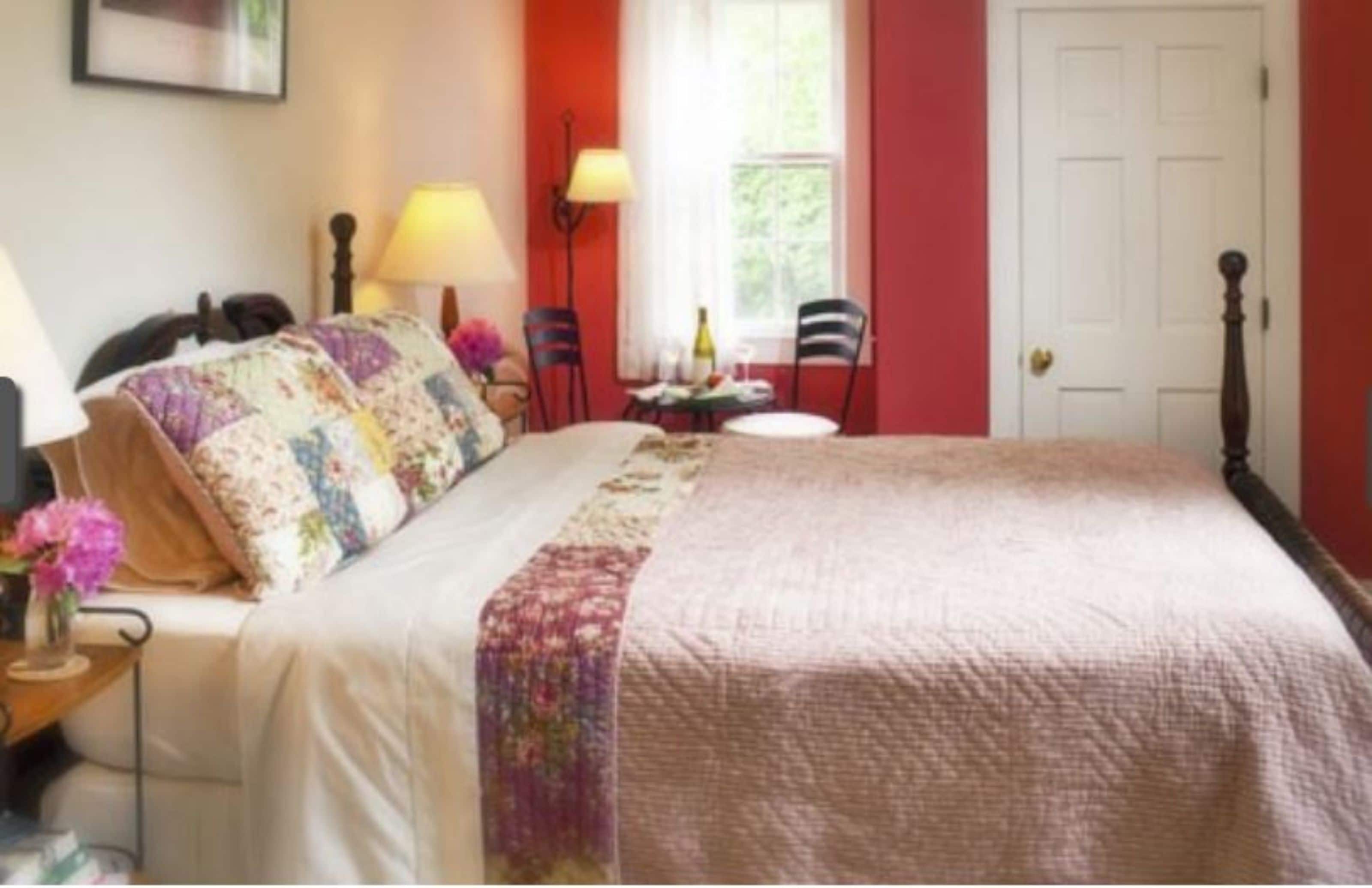 A neatly made bed with a floral bedspread and pillows, a bedside table with a lamp, and a chair near a window with a view of greenery.