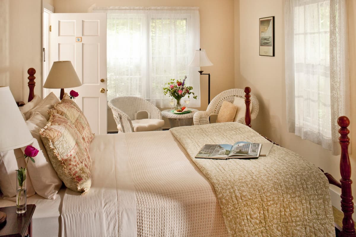 A neatly arranged bedroom with a bed, two chairs, a small table with a vase of flowers, and a lamp.