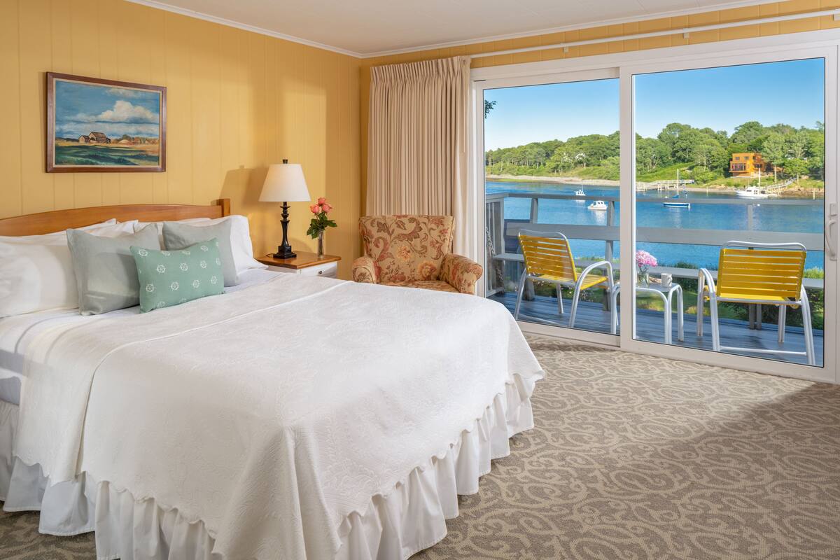 A hotel room with a large bed, a chair, a table, and a view of a lake with boats.