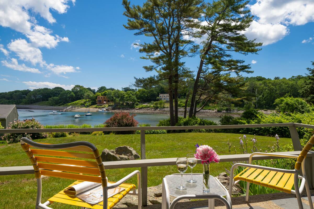 A terrace with yellow and white chairs, a table with a vase and wine glasses, overlooking a lake with boats and a bridge.