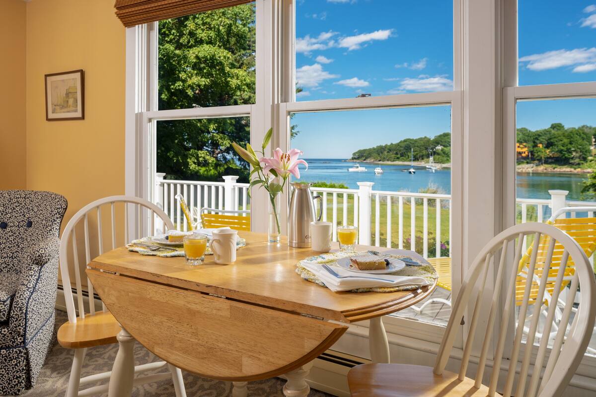 A dining area with a wooden table, white chairs, and a view of a lake with boats.
