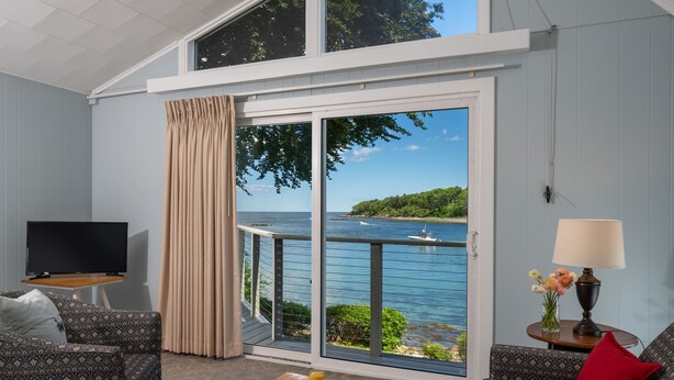 A room with a large sliding glass door leading to a balcony with a view of the sea. There is a TV on a stand, a dining table with breakfast items, and a lamp on a side table. - Dockside Guest Quarters & Restaurant (York)