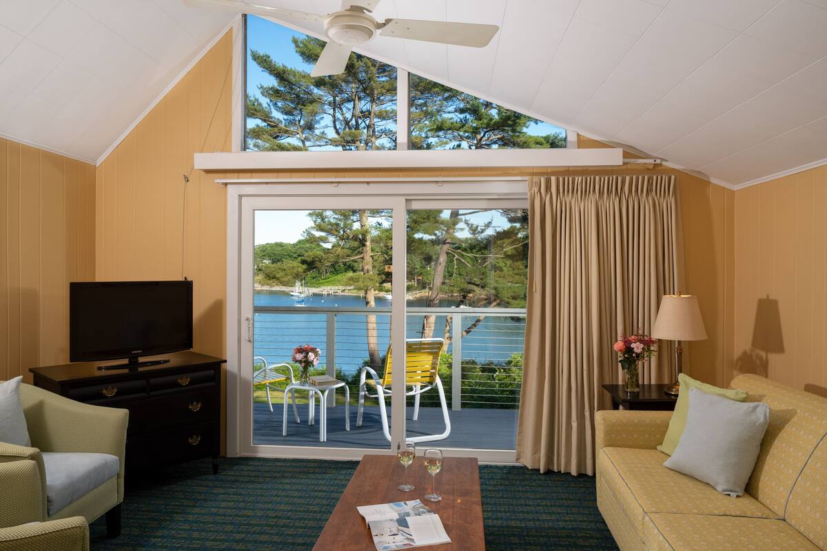 A room with a television, a sofa, a coffee table with a magazine, and a balcony with a view of a lake and trees.