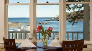 A dining table with a vase of pink flowers, two wine glasses, and a napkin, set against a backdrop of a scenic ocean view through large windows. - Dockside Guest Quarters & Restaurant (York)