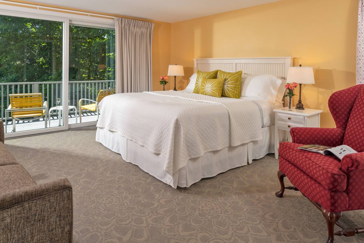 A hotel room with a large bed, two bedside tables, a red armchair, and a balcony with yellow chairs.