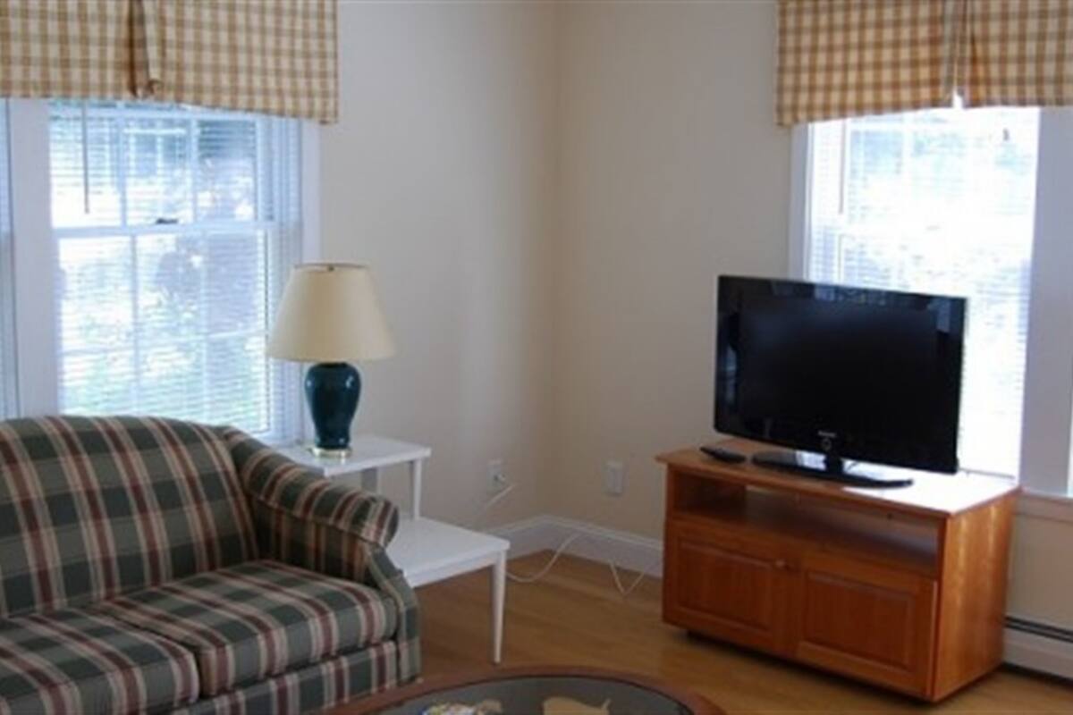 A living room with a plaid sofa, a television on a wooden stand, a white side table with a lamp, and a window with blinds.