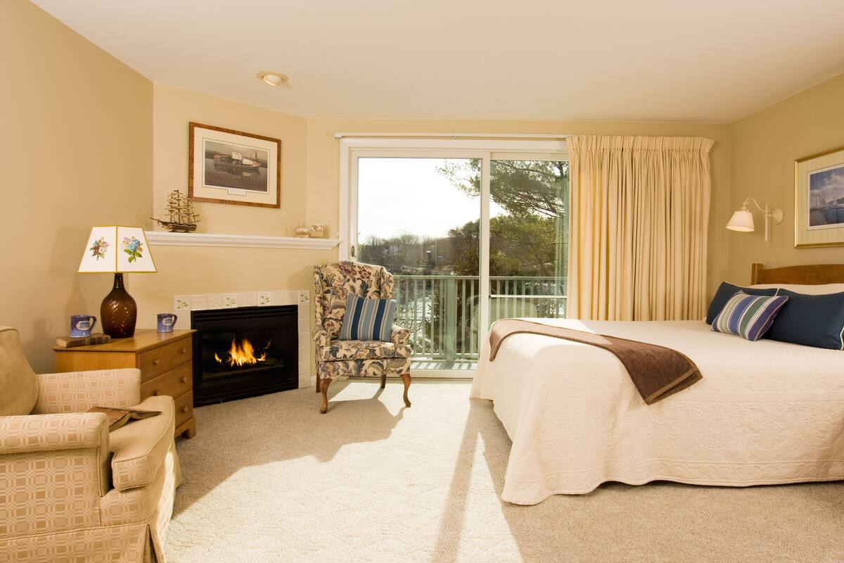 A bedroom with a fireplace, a large bed, a chair, and a view of the outdoors.