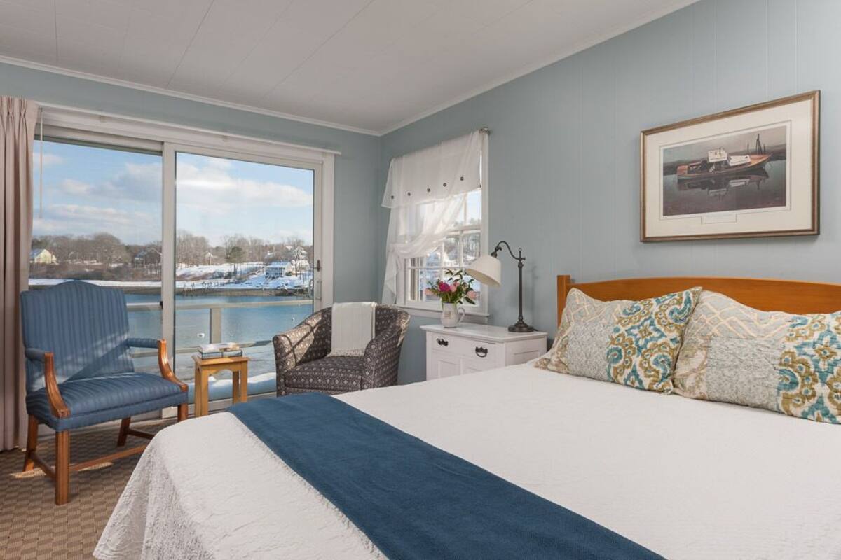A hotel room with a large bed, a chair, a small table with a lamp, and a window with a view of a marina.