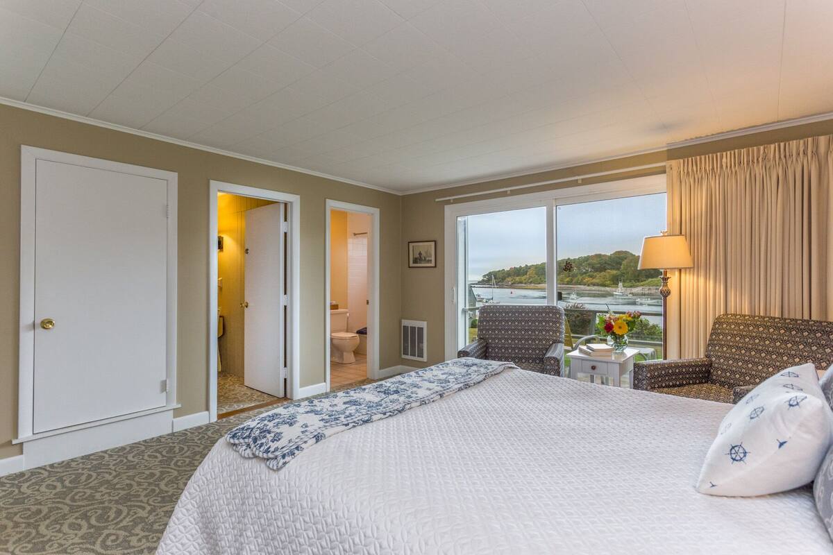 A hotel room with a large bed, a chair, a small table with a lamp, and a view of a river and trees.