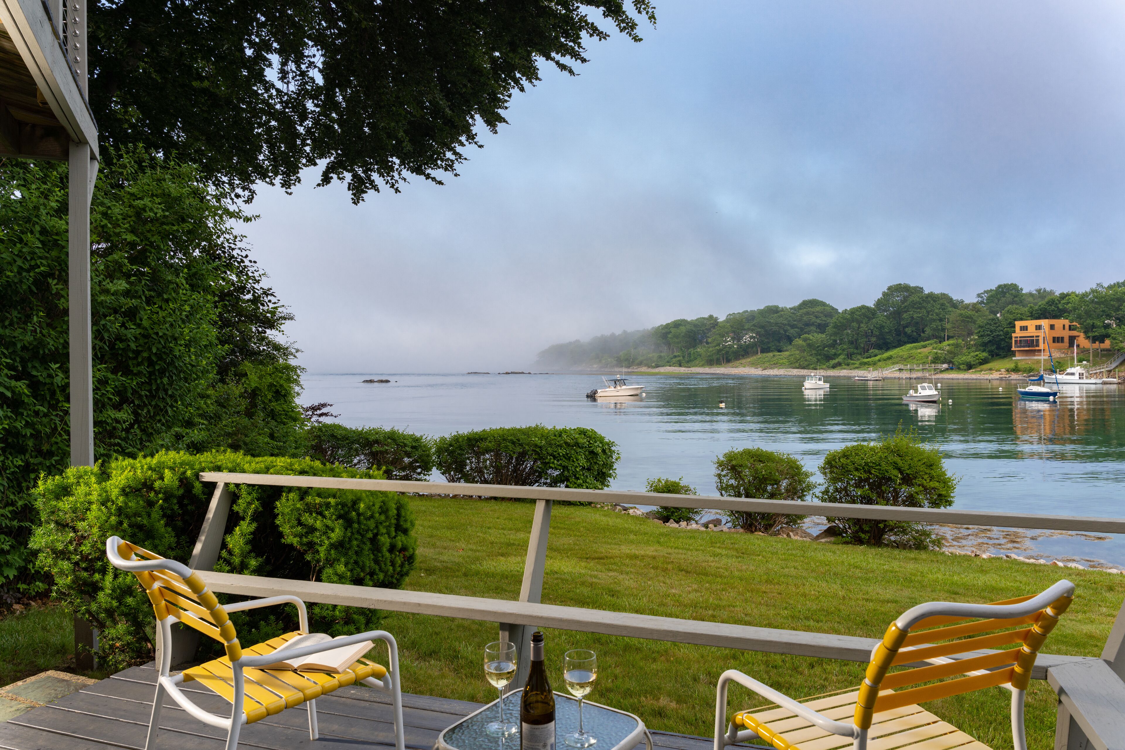 A deck with two chairs and a table, overlooking a calm body of water with boats.