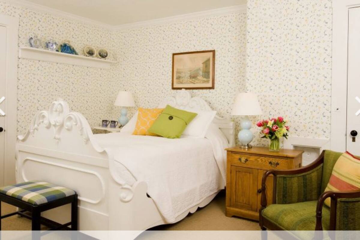 A bedroom with a white bed, a wooden nightstand, a green chair, and floral wallpaper.