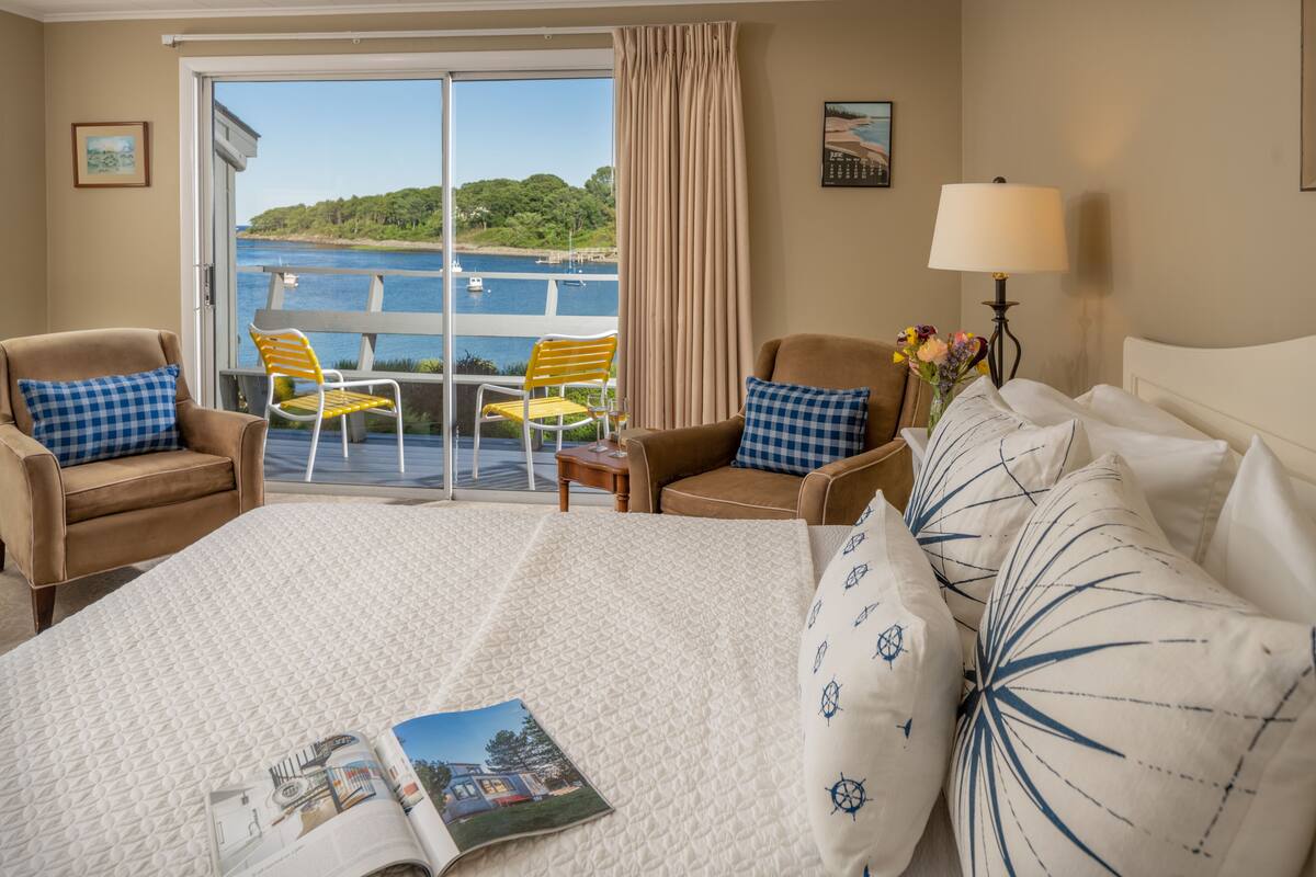A hotel room with a large bed, two armchairs, a small table with a lamp, and a view of a body of water through a sliding glass door.