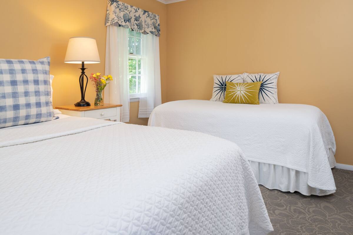 A hotel room with two beds, a nightstand with a lamp, a window with floral curtains, and a view of greenery outside.