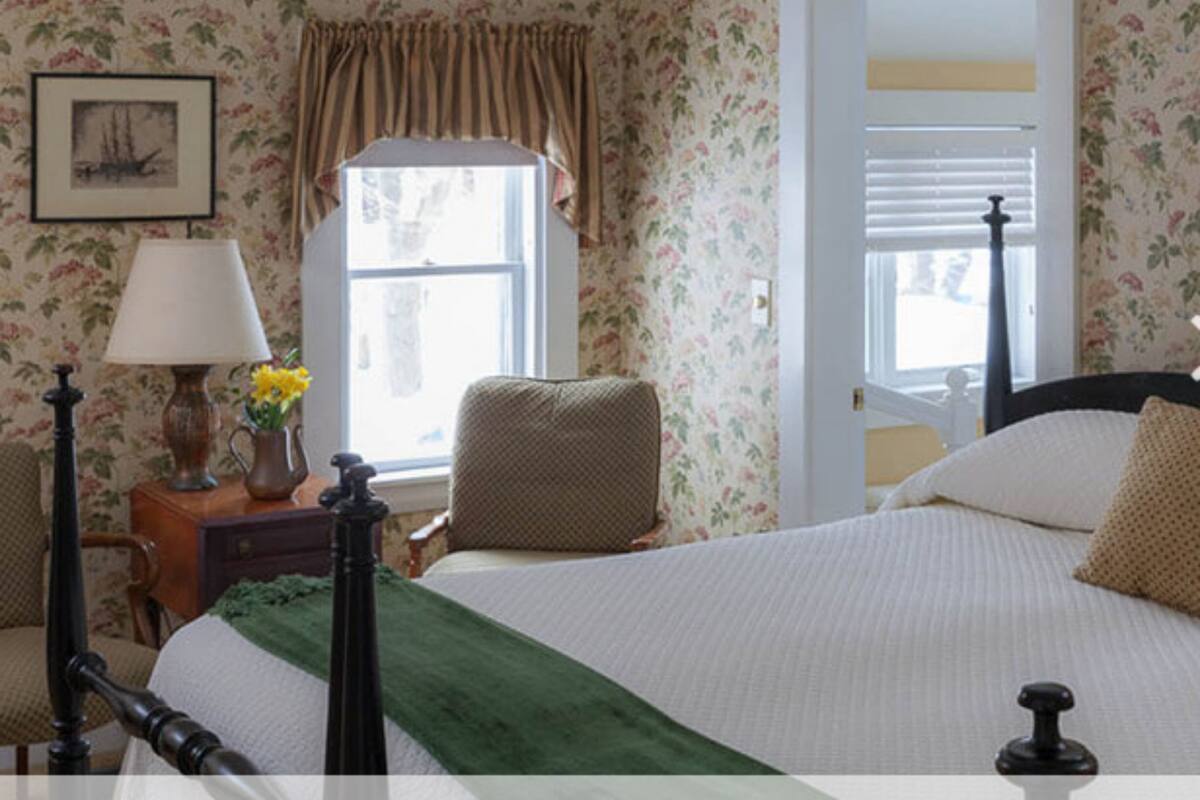 A bedroom with floral wallpaper, a bed with a green throw, a nightstand with a lamp and vase, and a window with curtains.