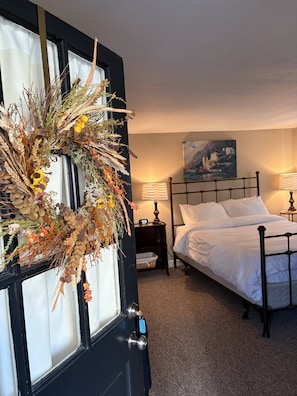 A bedroom with a bed, bedside tables, a nightstand, a lamp, a painting on the wall, and a wreath on a door.