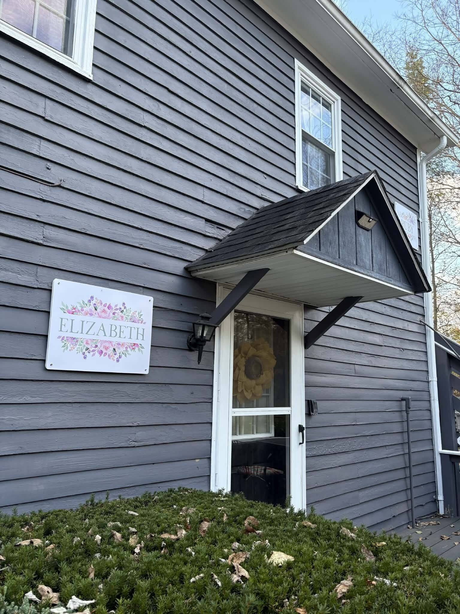 A house with a dark gray exterior, a white door, and a small porch. A sign with the name 'ELIZABETH' is visible on the wall.