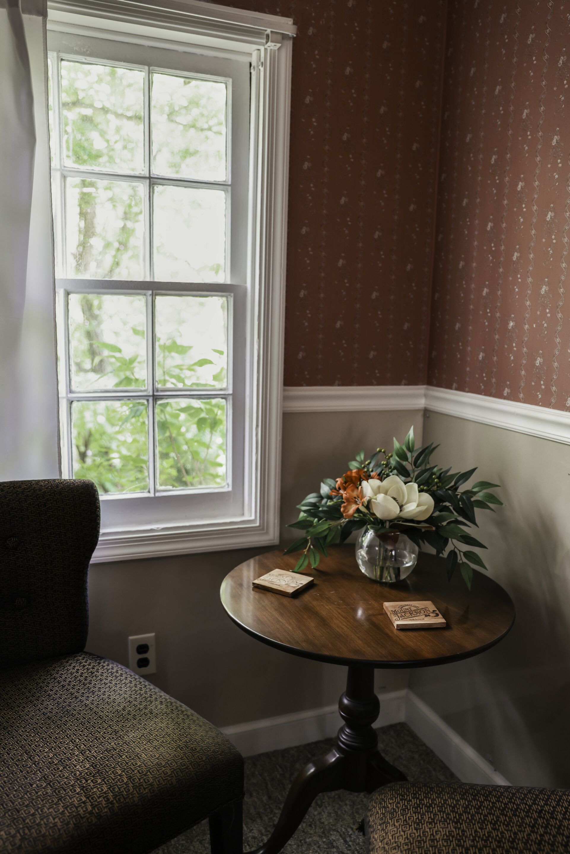 A corner room with a window, a round table with a vase of flowers, and patterned wallpaper.
