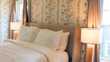 A neatly made bed with white linens, a wooden nightstand with a lamp, and floral wallpaper.