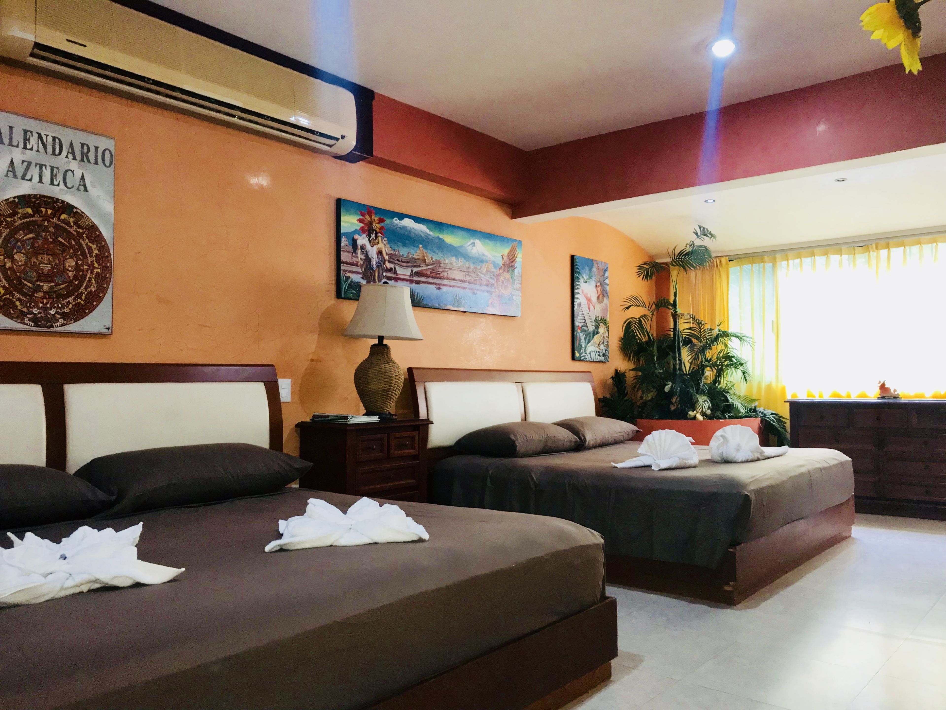 Quarto de hotel com duas camas, mesa de cabeceira, abajur e uma janela grande com cortinas.