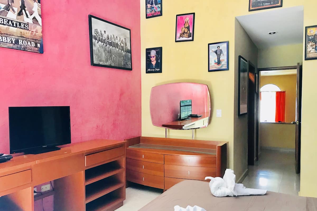 A hotel room with a pink wall, a wooden desk, a flat-screen TV, a bed with a white towel, and various framed pictures on the wall.