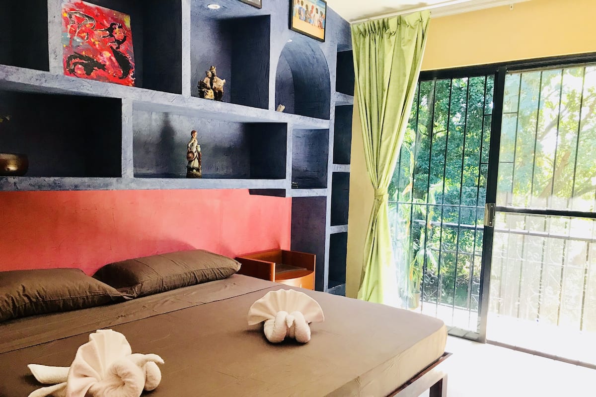 A room with a bed, pillows, a nightstand, a shelf with decorative items, and a view of greenery through a window.