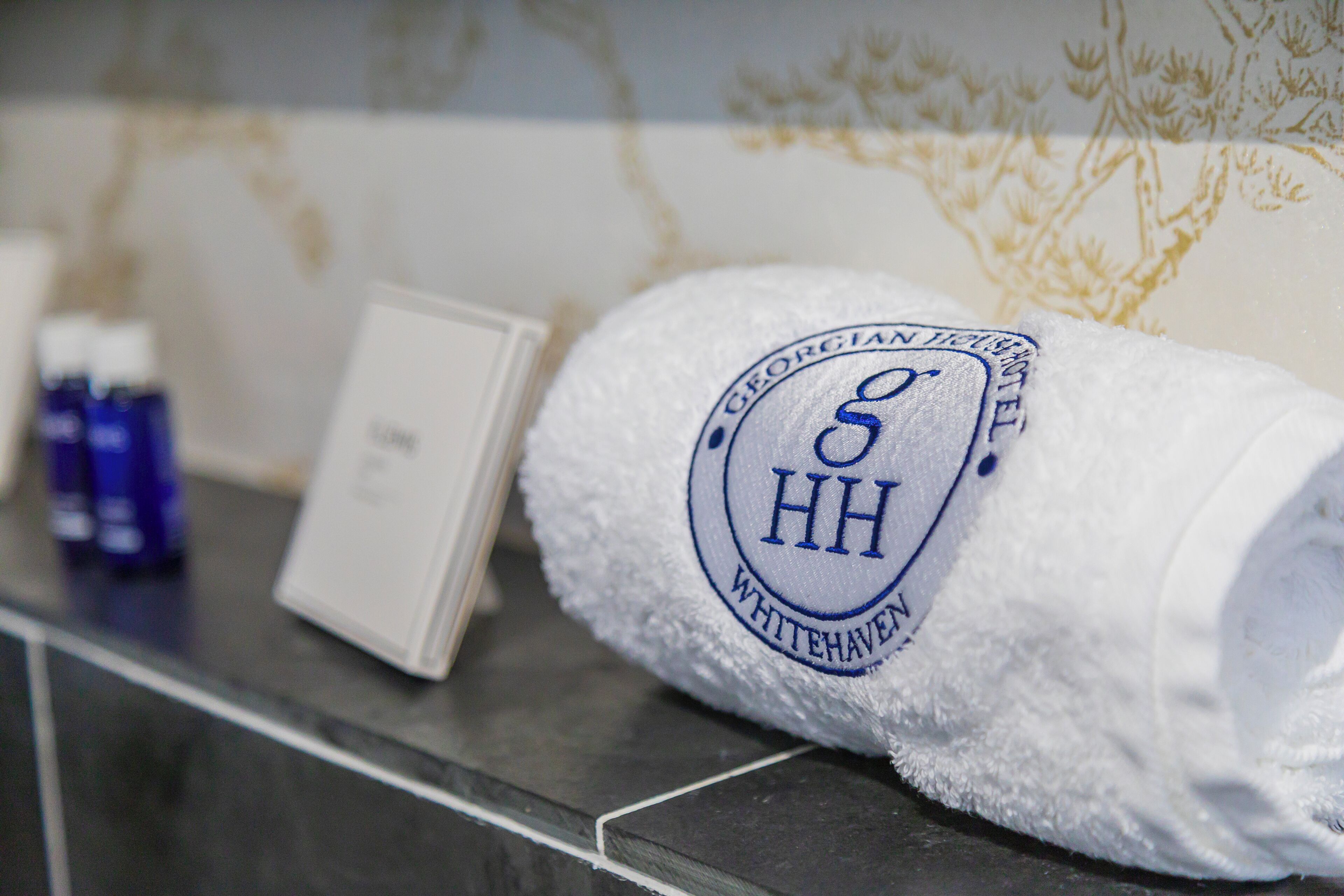 A white towel with a logo and text, placed on a tiled surface next to a small white card and two blue bottles.