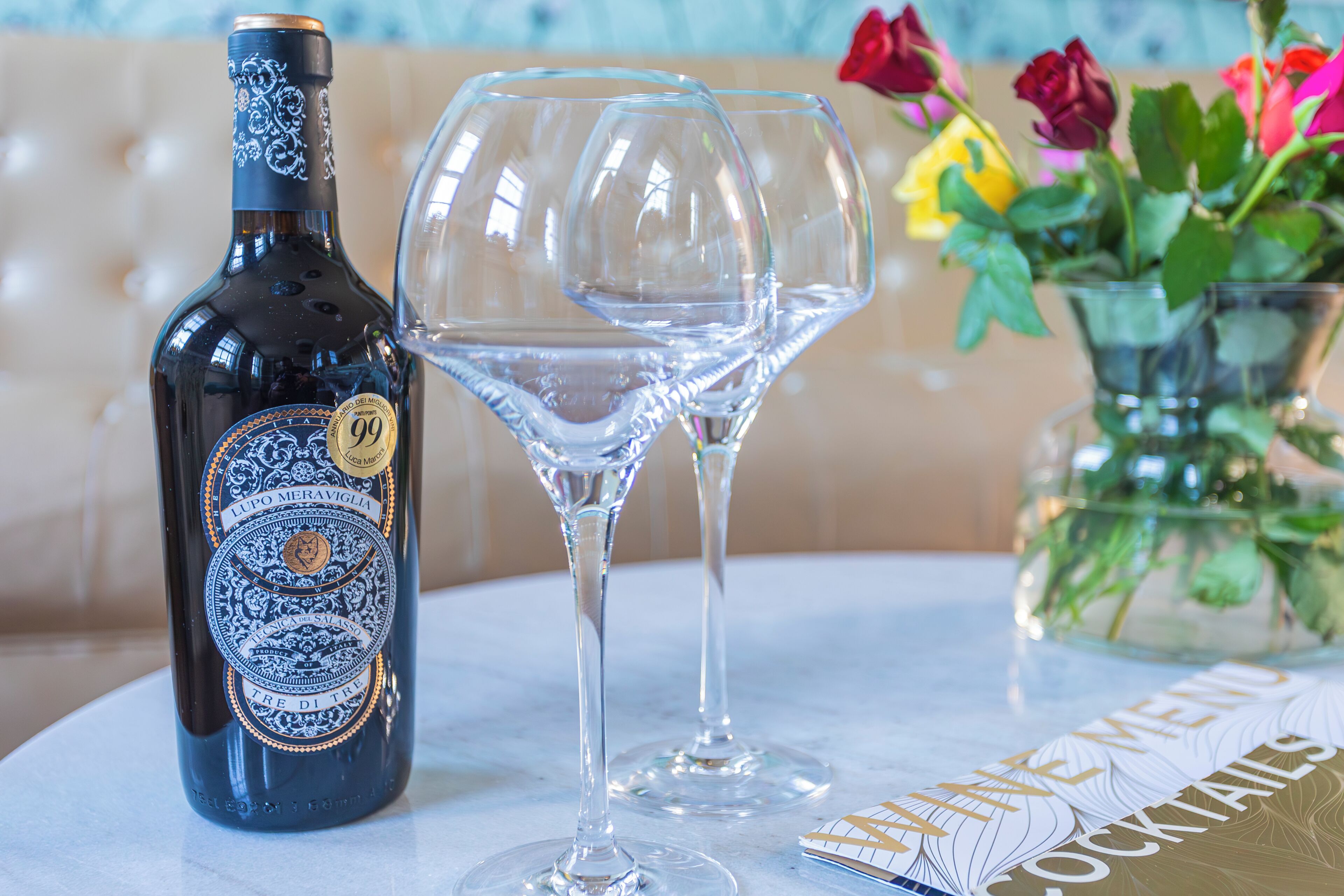 Una bottiglia di vino Lupo Meraviglia, due bicchieri da vino vuoti e un menu intitolato "The Menu Cocktails" su un tavolo.