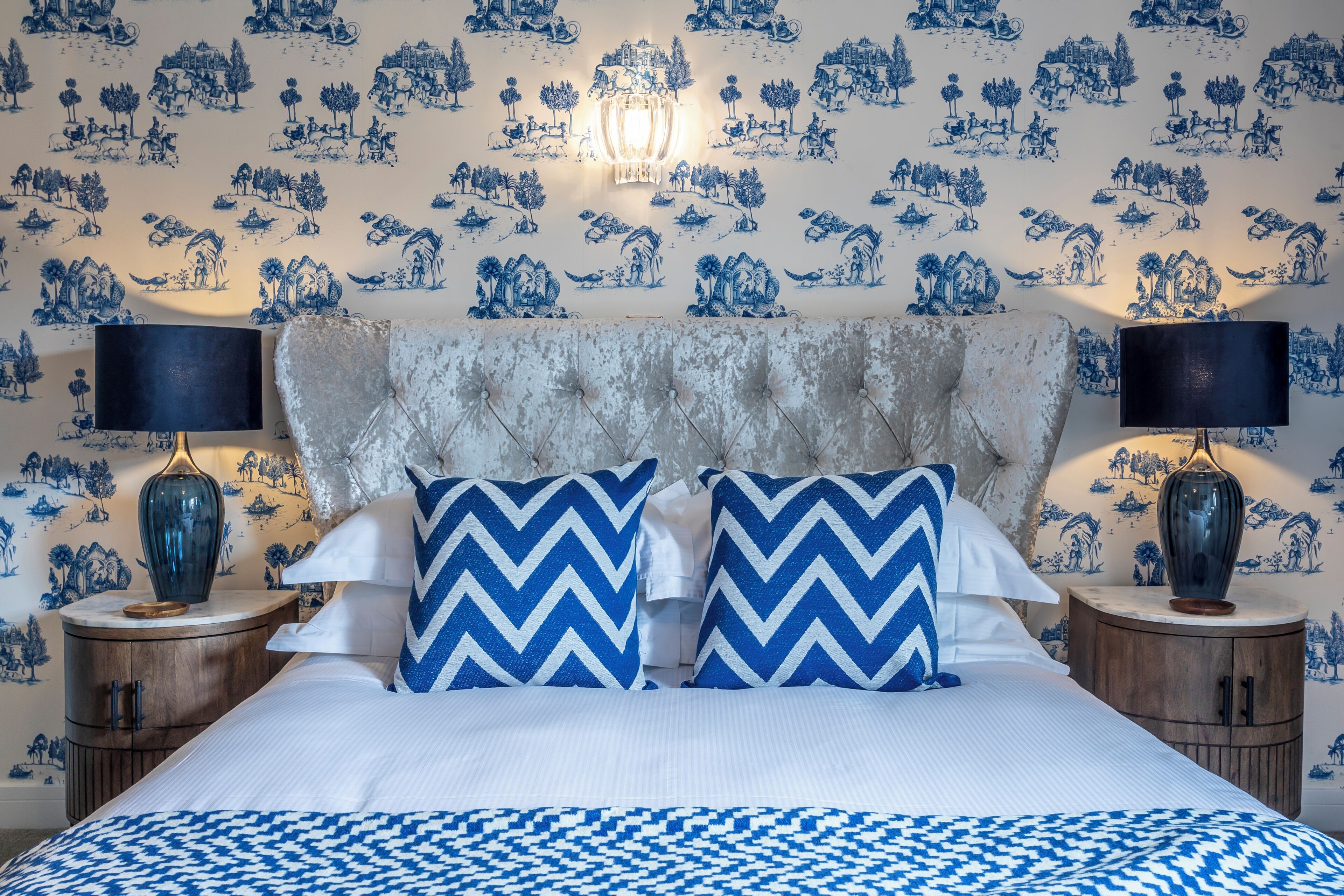 A bedroom with a bed, two nightstands, a chandelier, and a patterned wallpaper.