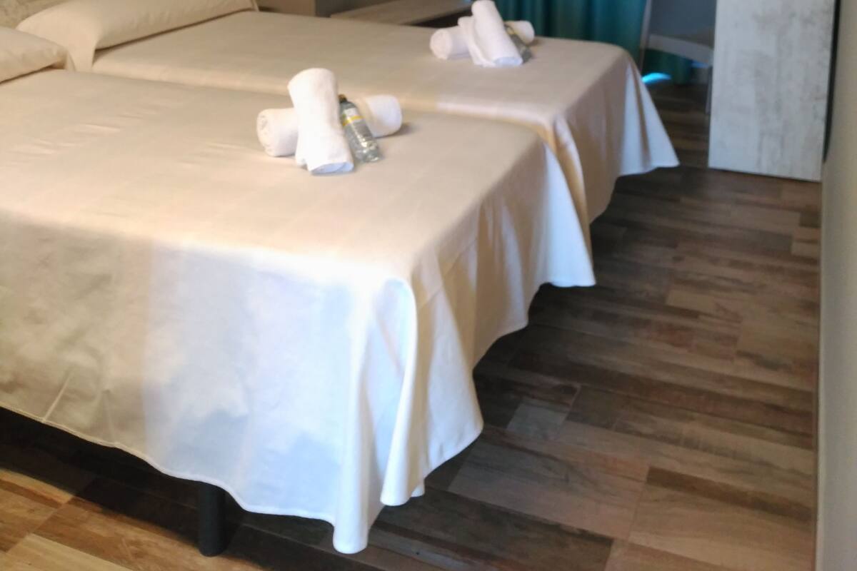 Une chambre d’hôtel avec deux lits, chacun étant équipé de draps et de serviettes blancs. Le sol est en bois et un rideau vert se trouve au fond de la pièce.