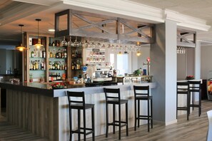 A bar area with a wooden bar counter, multiple stools, hanging light fixtures, and shelves stocked with bottles.