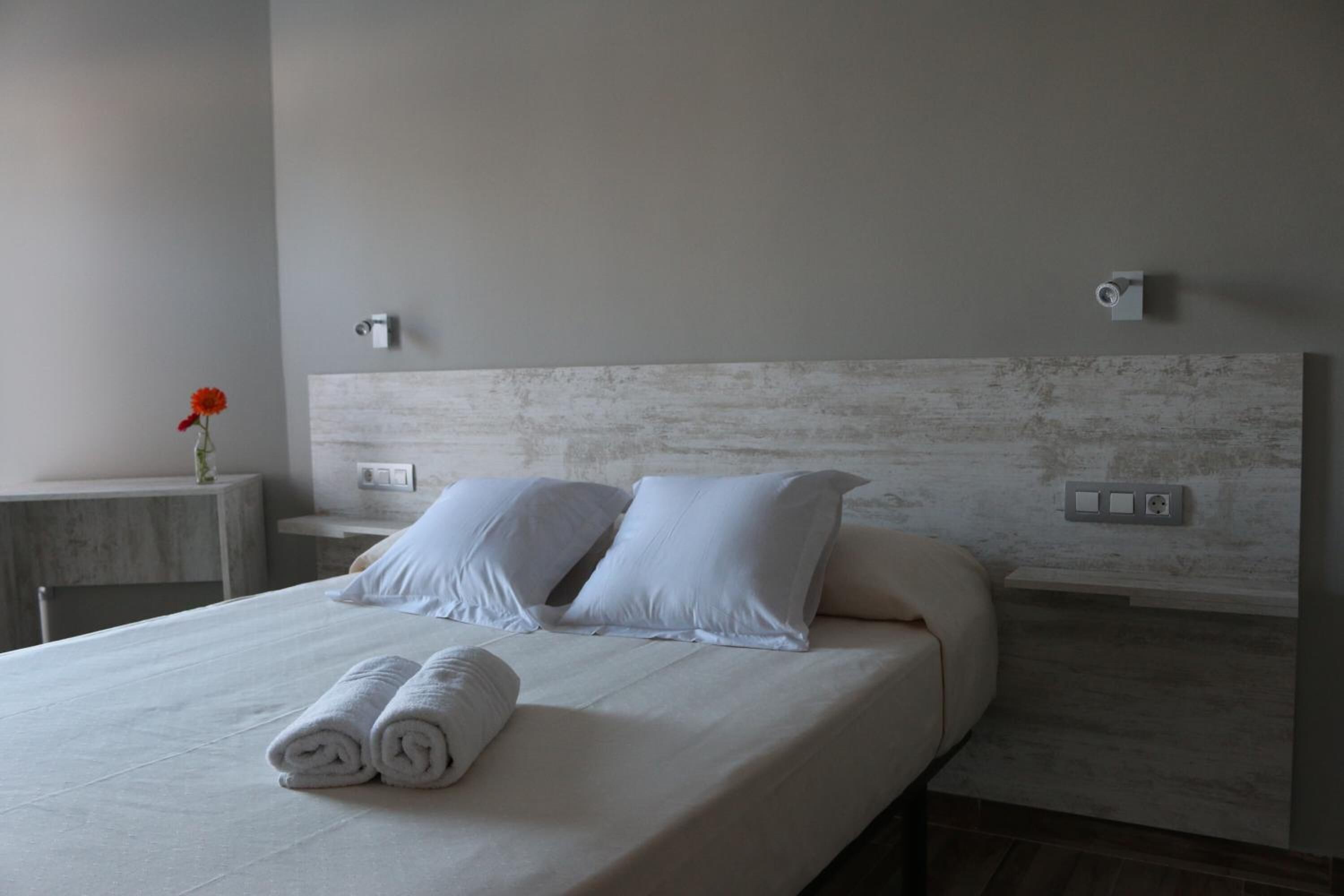 A neatly made bed with white linens and pillows, a small bedside table with a vase of flowers, and a modern headboard.