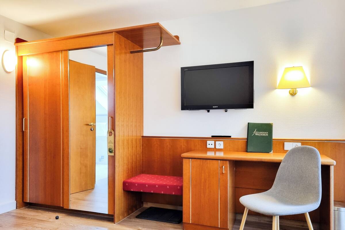 Een kleine kamer met een houten garderobe, een bureau met stoel, een televisie en een wandlamp.