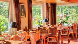 Un espace repas avec des chaises et des tables à motifs, de grandes fenêtres avec des rideaux et une vue sur la verdure à l’extérieur.