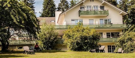A two-story building with a balcony, surrounded by trees and a grassy area with chairs.