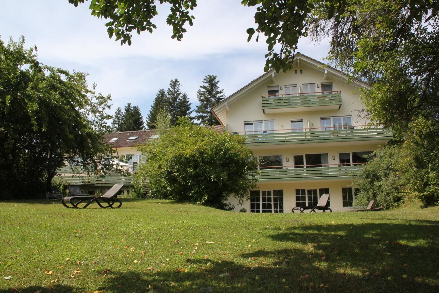 A two-story building with a balcony, surrounded by trees and a grassy lawn.