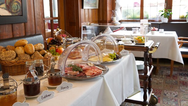 Una mesa de buffet con diversos alimentos y condimentos, como carnes, quesos y frutas, en una sala con paredes de madera y ventanas amplias.