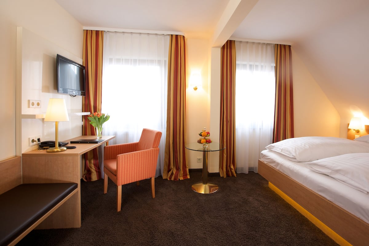 A hotel room with a bed, a desk with a lamp, a chair, a small table with fruit, and curtains.
