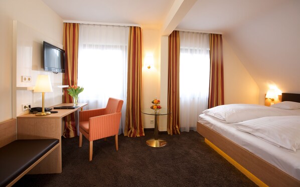 Une chambre d’hôtel avec un lit, un bureau doté d’une lampe, une chaise, une petite table avec des fruits et des rideaux.