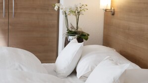 Una cama bien hecha con sábanas y almohadas blancas, un florero con flores blancas sobre una mesita de noche y un armario de madera con una luz empotrada en la pared.