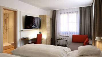 A hotel room with a bed, a desk, a TV, a shower, and a window with curtains.